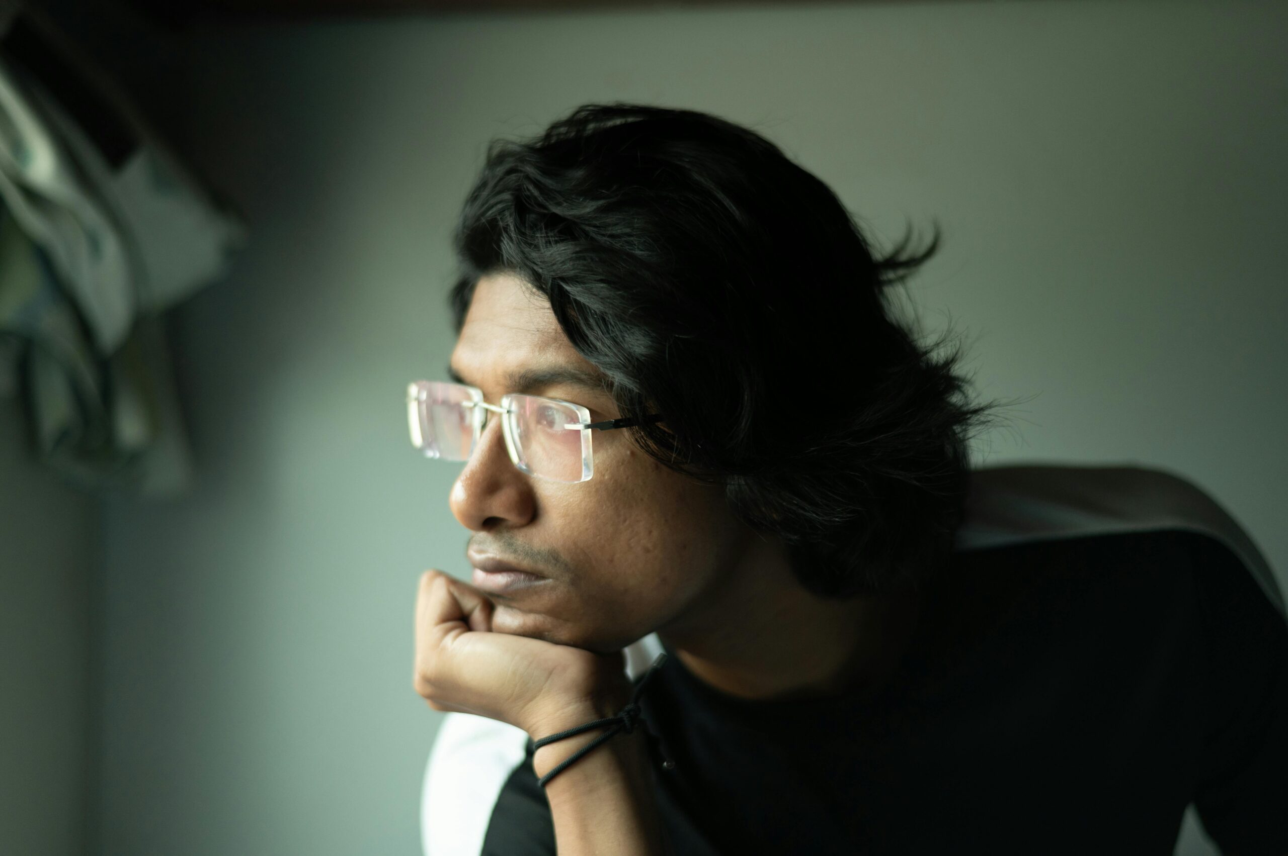 A young man wearing eyeglasses gazes thoughtfully out the window in a side profile view.